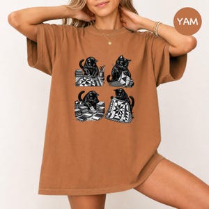 Può includere: Una t-shirt marrone con una stampa grafica di quattro gatti neri impegnati in attività di quilting. Le illustrazioni mostrano i gatti intenti a tagliare stoffa, cucire, organizzare pile di quadrati fantasia e reggere una trapunta finita.