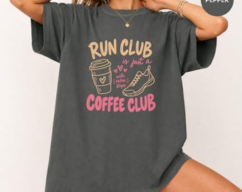 May include: A charcoal grey short-sleeve t-shirt featuring a graphic with the text RUN CLUB is just a COFFEE CLUB with extra steps. The design includes a line-art coffee cup, a running shoe, and small hearts in shades of pink and cream.
