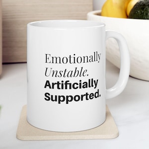 Peut inclure: Un mug en céramique blanche avec une anse en forme de C, portant l'inscription noire Emotionally Unstable. Artificially Supported. Le mug est posé sur un dessous de verre carré, présentant un design épuré pour la consommation de boissons.