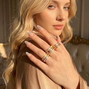 Può includere: Una collezione di anelli color oro indossati sulle dita, caratterizzati da vari design tra cui fasce incastonate con pietre chiare e scintillanti e texture in metallo intrecciato. Gli anelli sono lucidi e riflettono la luce con finitura metallica.