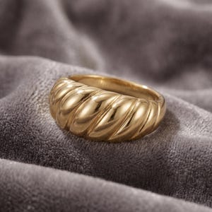 Könnte beinhalten: Ein goldfarbener Ring mit einem dicken, abgerundeten Band, das eine sich wiederholende diagonale Rippen- oder Verdrehungsstruktur aufweist. Die Metalloberfläche ist poliert und reflektierend, was Glanzlichter entlang der geschwungenen Kanten erzeugt.