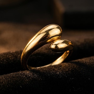 May include: A polished gold ring featuring an open bypass design with two smooth, rounded, teardrop-shaped ends that curve toward each other. The metal surface is highly reflective, catching the light to highlight its curved, fluid form.