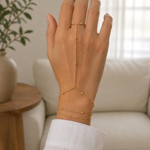 Könnte beinhalten: Eine goldfarbene Handkette mit einem zarten Gliederkettendesign und kleinen, gleichmäßig verteilten kugelförmigen Perlen. Das Schmuckstück verbindet einen am Mittelfinger getragenen Ring mit einem mehrlagigen Armband um das Handgelenk.