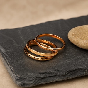 Puede incluir: Un conjunto de tres anillos de tono dorado apilados. Dos de los anillos presentan un acabado liso y pulido, mientras que el anillo central tiene una superficie texturizada y brillante. Los anillos son circulares con bandas delgadas.