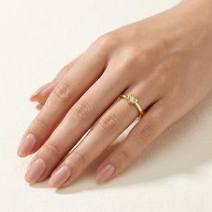 Puede incluir: Un anillo de oro con un pequeño engaste en forma de corazón que sostiene una sola piedra preciosa redonda y transparente. La banda es fina y pulida, mostrando un acabado metálico liso. El diseño de corazón está centrado en la banda.