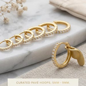 Puede incluir: Un conjunto de pendientes de aro dorados con piedras en pavé en tamaños graduados de 5 mm a 9 mm. Los aros tienen un cierre de bisagra y se muestran en fila, con un aro separado. El texto dice: CURATED PAVÉ HOOPS, 5MM - 9MM.