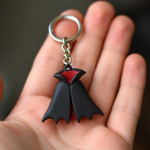 May include: A keychain featuring a miniature black cape with a red interior lining. The cape is attached to a silver-colored metal split ring by a short chain. The design is smooth and matte, depicting a stylized vampire-style cloak accessory.