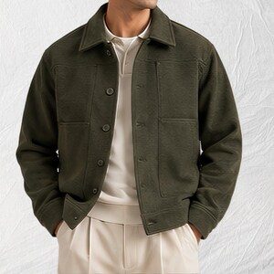 Könnte beinhalten: Eine olivgrüne Jacke mit Knopfleiste, strukturiertem Kragen, langen Ärmeln und zwei großen aufgesetzten Vordertaschen. Die Jacke wird über einem cremefarbenen Poloshirt und einer hellen Hose getragen, was für ein gepflegtes, geschichtetes Aussehen sorgt.