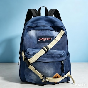 Könnte beinhalten: Ein blauer Rucksack in Denim-Optik mit einem JanSport-Logo-Aufnäher auf der Vorderseite. Der Rucksack verfügt über eine Reißverschlusstasche vorne, schwarze Schultergurte und ein beiges, verstellbares Gurtsystem mit schwarzen Kunststoffschnallen.
