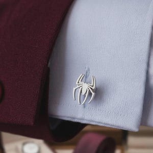 May include: A silver-toned metal cufflink shaped like a stylised spider with eight legs, attached to the cuff of a light blue dress shirt. The shirt cuff is partially covered by the sleeve of a dark burgundy blazer with a visible matching button.