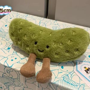 Op de afbeelding: Een groen pluche augurk-speeltje met een zachte, pluizige textuur, geborduurde gelaatstrekken en twee bruine ribfluwelen beentjes. Het speeltje is 15 cm groot en heeft een wit Jellycat-merklabel met een zwart kattenlogo en ananasicoon.