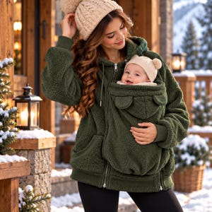 Könnte beinhalten: Ein dunkelolivgrüner Kapuzenpullover mit Reißverschluss aus flauschigem Sherpa-Fleece. Der Pullover verfügt über eine integrierte Fronttasche für ein Kleinkind, die mit kleinen dekorativen Bärenohren an der Taschenklappe versehen ist. Das Kleidungsstück hat lange Ärmel.
