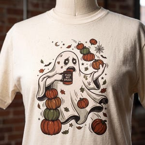 May include: A cream-colored t-shirt featuring a graphic of a white ghost holding a mug that reads PUGS NOT DRUGS. The ghost is surrounded by orange and green pumpkins, falling autumn leaves, a spider web, stars, and small bats.