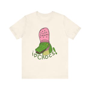 Könnte beinhalten: Ein cremefarbenes Kurzarm-T-Shirt mit einer Grafik eines grünen Alligators, der einen rosa Clog auf dem Kopf trägt. Unter dem Alligator steht der Text I heart CROCS! in grüner Schrift, wobei ein Herzsymbol das Wort love ersetzt.