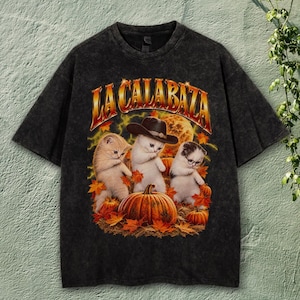 May include: A charcoal grey acid-wash t-shirt featuring a graphic print of three kittens among pumpkins and autumn leaves. The center kitten wears a cowboy hat, and the right kitten wears glasses. The text LA CALABAZA appears in gold above.