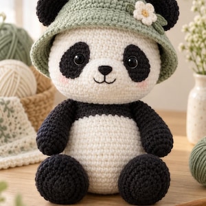 May include: A crocheted panda stuffed animal featuring a white and black body, black ears, and a sage green bucket hat adorned with a small white flower. The panda has black eye patches, a black nose, a stitched mouth, and soft pink blush on its cheeks.