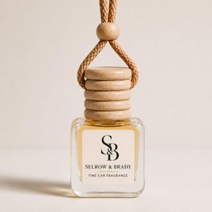 May include: A square glass car fragrance bottle with a light amber liquid, topped with a stack of five light-colored wooden rings and a braided hanging cord. The white label features the black text S&B, SELROW & BRADY, and FINE CAR FRAGRANCE.