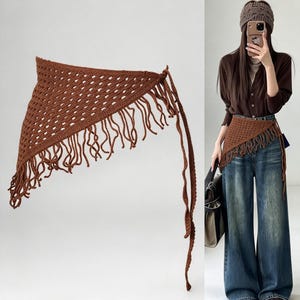 May include: A brown triangular crochet wrap featuring an open-knit diamond pattern and long fringe along the bottom edge. The accessory includes thin crocheted ties on the sides for fastening, showcasing a textured, handmade yarn construction.
