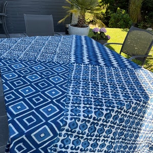 Op de afbeelding: Een blauw-wit patroon tafelkleed bedekt een tafel. Het tafelkleed is verdeeld in vier secties, elk met een ander geometrisch ontwerp. De patronen omvatten ruiten, zigzags en andere abstracte vormen. De tafel staat buiten.