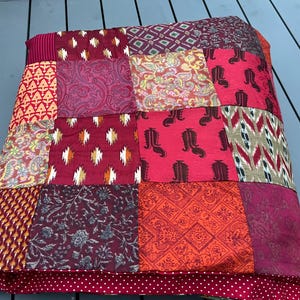 Peut inclure: Un coussin patchwork carré aux tons rouges, bordeaux et orange. Il est composé de divers tissus à motifs, notamment des motifs cachemire, géométriques et floraux. Bordure à pois rouges et blancs. Dimensions d'environ 40 cm x 40 cm.