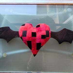 Puede incluir: Un cojín de peluche con forma de corazón y un patrón pixelado en tonos rojos y negros. El corazón tiene dos alas negras similares a las de un murciélago a los lados, con costuras que definen la forma y estructura de las alas.