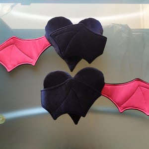 May include: Two plush heart-shaped ornaments featuring dark purple velvet bodies and bright pink bat wings. Each heart has a pointed bottom and stitched wing details, with the wings extending outward from the sides of the plush fabric hearts.