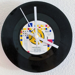 May include: A wall clock crafted from a black vinyl record featuring a central label with a colorful grid pattern. The label displays the text Tarzan Boy by Baltimora, Manhattan Records, and 1985. It has three plain white clock hands.