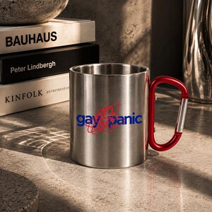 Puede incluir: Una taza de acero inoxidable de color plateado con un asa de mosquetón roja. La parte frontal de la taza muestra el texto "gay panic" en letras minúsculas azules, superpuesto con un diseño gráfico circular de garabatos rojos.