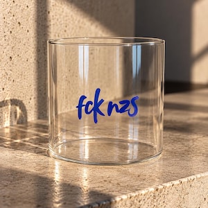 Könnte beinhalten: Ein zylindrisches, klares Glas mit dem Schriftzug fck nzs, der in einer kräftigen, handschriftlichen blauen Schrift in der Mitte aufgedruckt ist. Das Glas hat eine glatte, transparente Oberfläche und ein schlichtes Design.