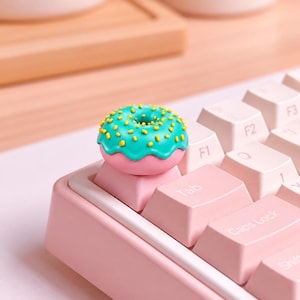 Puede incluir: Un teclado mecánico rosa con una tecla personalizada en forma de dónut sobre la tecla Tab. El dónut tiene glaseado color verde azulado y virutas amarillas. El teclado tiene teclas rosa claro con letras blancas, incluyendo F1, F2, F3, Tab y Caps Lock.