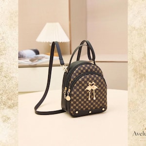 Puede incluir: Una mochila pequeña de color marrón y negro con un patrón geométrico repetitivo, un asa superior y correas ajustables. El bolsillo frontal está adornado con un emblema de metal dorado en forma de cisne con cadenas colgantes y tres tachuelas.