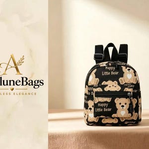 Op de afbeelding: Een zwarte mini-rugzak met een herhalend patroon van beige teddyberen met witte hartvormige buikjes. De tas heeft een voorvak met rits, zwarte verstelbare schouderbanden en de tekst Happy Little Bear op de stof gedrukt.