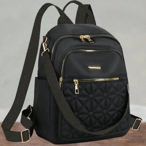 Puede incluir: Una mochila negra con un bolsillo frontal acolchado de patrón geométrico, cremalleras metálicas doradas y correas ajustables. La bolsa incluye un asa superior, un compartimento principal con cremallera y un bolsillo frontal más pequeño con placa dorada.