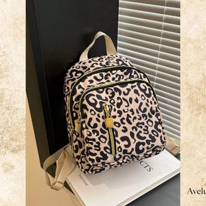 May include: A small backpack featuring a beige and black leopard print pattern. The bag includes a top handle, adjustable beige straps, and a front zippered pocket with a gold-toned rectangular zipper pull. The main compartment also has a zipper.