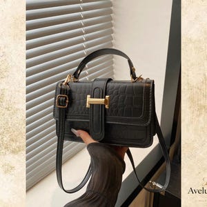 May include: A black rectangular handbag with a textured crocodile-embossed pattern, featuring a top handle and a detachable shoulder strap. The front flap is secured with a vertical strap and a gold-toned metal H-shaped clasp hardware.
