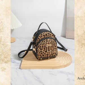 Könnte beinhalten: Ein kleiner Rucksack mit einer Basis in schwarzer Lederoptik und einer Vorderseite mit Leopardenmuster. Der Rucksack verfügt über einen Tragegriff, verstellbare Schultergurte, goldfarbene Beschläge und einen dekorativen goldenen Flamingo-Anhänger.