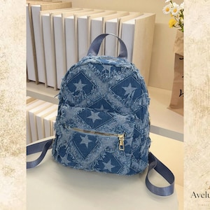 May include: A blue denim backpack featuring a repeating star pattern and frayed edges. The bag includes a top handle, two shoulder straps, and a front zippered pocket with a gold-toned metal zipper pull. The overall construction is structured.