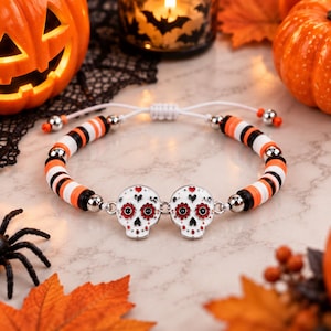 Op de afbeelding: Een armband met twee witte suikerschedelbedels met rode en zwarte bloemendetails, verbonden door zilverkleurige metalen schakels. De band bestaat uit afwisselende oranje, zwarte en witte platte ronde kralen, zilverkleurige tussenstukjes en een wit koord.