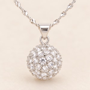 May include: A silver-toned necklace featuring a spherical pendant encrusted with numerous clear, round-cut stones. The pendant hangs from a delicate, textured silver-toned chain that connects to a polished, triangular-shaped bail at the top.