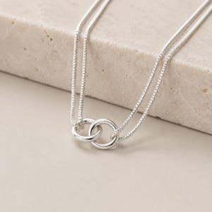 May include: A silver-toned necklace featuring two interlocking circular rings as a central pendant. The chain is a fine, textured snake-style chain that loops through both rings, creating a double-strand effect with a polished metallic finish.
