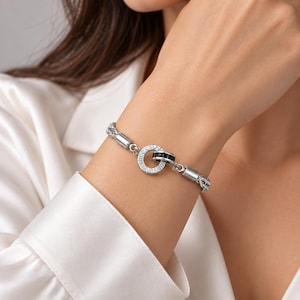 Puede incluir: Una pulsera de cadena en tono plateado con un eslabón circular central adornado con cristales transparentes y un anillo entrelazado más pequeño con piedras negras. La pulsera está construida con segmentos metálicos cilíndricos pulidos y una cadena tejida.