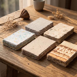Puede incluir: Cinco pilas rectangulares de papel hecho a mano con bordes irregulares. Una pila presenta motas de colores, otra tiene una textura acanalada, otra contiene elementos botánicos, otra es beige lisa y otra muestra un patrón tie-dye marrón.