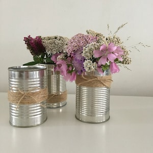 May include: Three silver cans with a rustic twine bow around the top, holding a bouquet of pink and white flowers.