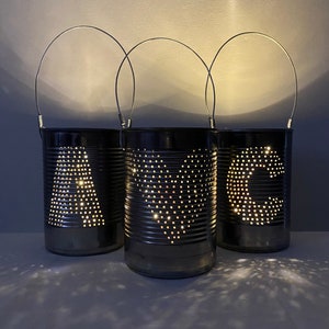 May include: Three handmade tin can lanterns with wire handles. The lanterns are dark grey with the letters "A", "C", and a heart shape punched out, allowing light to shine through. The lanterns are illuminated from within, casting a warm glow.