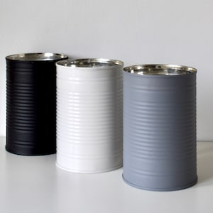 Könnte beinhalten: Drei leere Metalldosen, eine schwarze, eine weiße und eine graue, mit silbernen Deckeln. Die Dosen stehen in einer Reihe auf einer weißen Oberfläche.