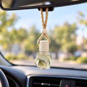 May include: A clear, faceted glass car air freshener bottle containing a light yellow liquid. The bottle has a ribbed wooden cap and a beige braided cord with gold metallic threading, suspended from a vehicle rearview mirror.