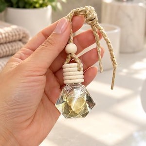 May include: A small, clear, diamond-shaped glass bottle for fragrance, featuring a faceted design. It includes a light-coloured wooden cap with horizontal ridges, a round wooden bead, and a braided beige cord with gold metallic threading.