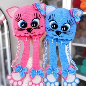 Peut inclure: Deux bonnets chat au crochet, un rose et un bleu, avec de grands yeux brodés à longs cils, des nez noirs et des moustaches. Chaque bonnet possède des oreillettes avec des tresses terminées par des moufles en forme de patte à nœuds bleus.