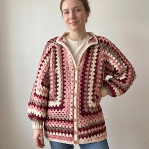 Peut inclure: Un cardigan en crochet à manches longues avec un motif de carrés granny dans des tons bordeaux, vieux rose, rose clair et crème. Le vêtement présente un col, une fermeture boutonnée avec des boutons marron clair et des poignets côtelés.