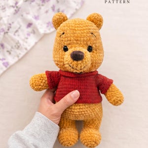 Peut inclure: Un ours en peluche crocheté jaune doré portant un pull rouge. L'ours a des yeux en perles noires, un nez marron foncé et des sourcils brodés. L'image inclut le texte PDF crochet pattern en haut et quatre petites icônes de drapeaux en bas.